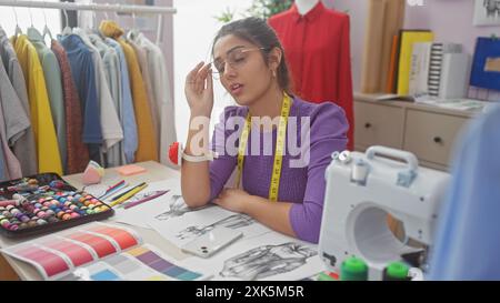 Eine nachdenkliche junge hispanische Frau in einem bunten Atelier, umgeben von Mode-Design-Tools und Kleidungsstücken. Stockfoto