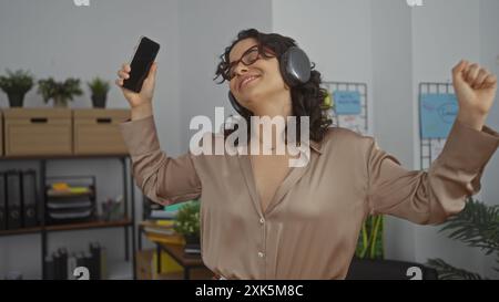 Eine junge brünette hispanische Frau, die Musik mit Kopfhörern genießt, während sie fröhlich in einer Innenraumbüros tanzt. Stockfoto