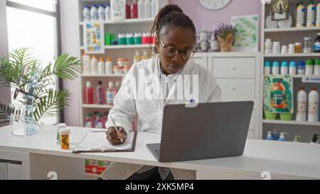 Frau, die in einer Apotheke arbeitet, einen Labormantel und eine Brille trägt, einen Laptop benutzt und Notizen in einer Drogerie im Innenbereich macht, umgeben von Regalen mit p Stockfoto