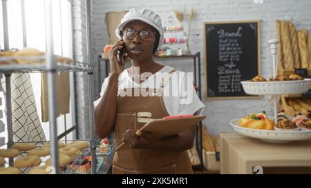 Junge afrikanerin mit Brille und weißem Hut in einer Bäckerei, die am Telefon spricht, während sie ein Klemmbrett hält, umgeben von Gebäck, Brot und Bak Stockfoto