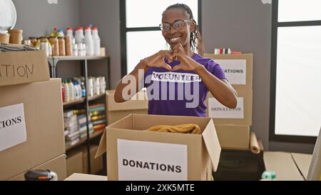 Eine lächelnde afroamerikanerin macht mit ihren Händen ein Herzzeichen, trägt ein freiwilliges T-Shirt in einem Spendenzentrum. Stockfoto