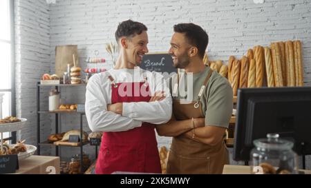 Zwei Erwachsene Männer in einer Bäckerei mit überkreuzten Armen, Schürzen, lächeln sich an und zeigen frisch gebackenes Brot im Hintergrund. Stockfoto