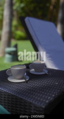 Zwei Keramikbecher Kaffee auf einem Korbtisch mit üppigem Grün im Hintergrund bedeuten eine ruhige Atmosphäre im Freien. Stockfoto