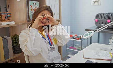 Attraktive blonde Frau in einem weißen Mantel bildet mit ihren Händen eine Herzform, während sie in einem Tierklinikzimmer lächelt. Stockfoto