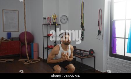 Junger Mann, der in einem Fitnessstudio mit Geräten um ihn herum trainiert, konzentriert sich intensiv auf ein Training in einem gut beleuchteten Raum mit Trophäen und Trainingsausrüstung im bac Stockfoto