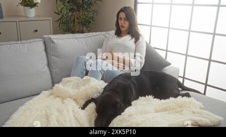 Eine junge hispanische Frau mit Magenschmerzen sitzt auf einer Couch mit einem schwarzen labrador-Hund in einem städtischen Innenraum. Stockfoto