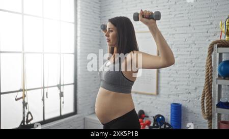 Eine junge hispanische schwangere Frau, die in einem Fitnessstudio Gewichte hob und aktive Mutterschaft in einem Sportzentrum präsentiert. Stockfoto