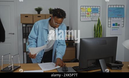 Junger afroamerikanischer Mann, der in einem Büro arbeitete, konzentrierte sich auf die Prüfung von Papieren an seinem Schreibtisch mit einem Computer und Organigrammen im Hintergrund. Stockfoto
