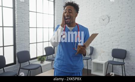 Ein hübscher junger afroamerikaner in blauen Schutten hält ein Klemmbrett und lacht im Wartezimmer des Krankenhauses. Stockfoto