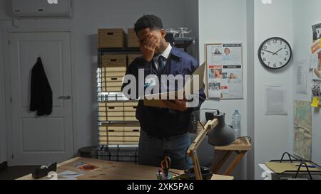 Ein afroamerikanischer Detective erscheint gestresst in einem überfüllten Polizeibüro und hält eine Akte mit Beweismitteln neben einer Tafel. Stockfoto
