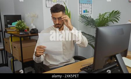 Gutaussehender hispanischer Mann, der Dokumente an seinem Schreibtisch im Innenbereich liest, umgeben von Pflanzen und Arbeitsmaterialien, konzentriert und mit Büroaufgaben beschäftigt. Stockfoto