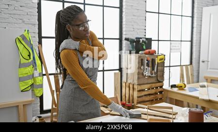 Eine schwarze Frau mit Zöpfen, die Rückenschmerzen in einer Tischlerei in Innenräumen hat, mit Schutzbrille und lässiger Kleidung. Stockfoto