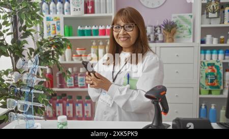 hispanische Frau mittleren Alters, die in einer Apotheke arbeitet, lächelt, während sie ein Smartphone benutzt, umgeben von verschiedenen Produkten und einem Strichcode-Scanner in einem bunten Stockfoto