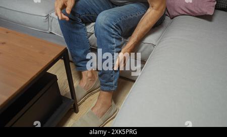 hispanischer Mann mittleren Alters, der im Wohnzimmer sitzt und sein Bein berührt, während er Jeans und Hausschuhe trägt Stockfoto