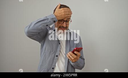 Reifer hispanischer Mann, der überrascht ist, während er auf ein Smartphone über isoliertem weißen Hintergrund blickt Stockfoto