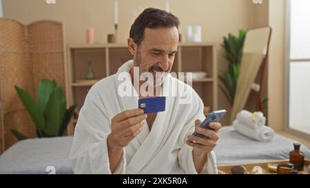 Hispanischer Mann in einem Spa-Zimmer, der einen weißen Bademantel trägt, eine Kreditkarte in der einen Hand und ein Smartphone in der anderen Hand hält. Stockfoto