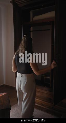 Eine junge Frau öffnet einen hölzernen Kleiderschrank in einem sanft beleuchteten Innenraum, was ein Gefühl von Zuhause und Neugier weckt. Stockfoto