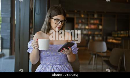 Schöne hispanische Frau, lässig in Gläsern gekleidet, genießt eine heiße Tasse Kaffee an einem Kaffeetisch, benutzt ihr Telefon und sonnt sich in der Innenentspannung im Stockfoto