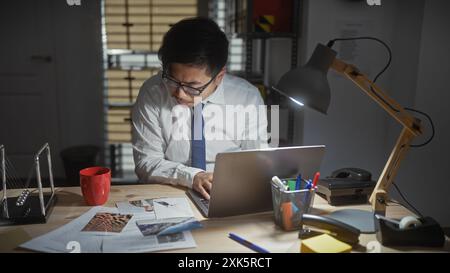 Fokussierter asiatischer Mann mit Brille, der spät nachts in einem Büro mit Laptop, Dokumenten und Schreibtischlampe arbeitet. Stockfoto