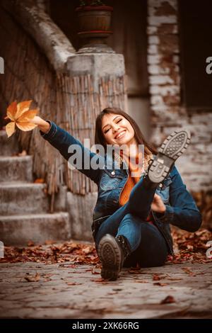 Eine Frau in einer Jeansjacke sitzt auf dem Boden vor Steintreppen, lächelt und hält ein gefallenes Blatt. Stockfoto