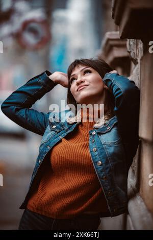 Eine junge Frau mit kurzen dunklen Haaren lehnt sich an eine Mauer in einer Stadt. Sie trägt eine blaue Jeansjacke über einem orangefarbenen Pullover. Sie schaut mit einem du auf Stockfoto