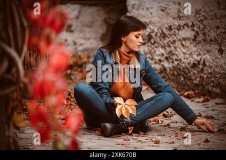 Eine Frau mit Jeansjacke und orangefarbenem Pullover sitzt auf dem Boden in einer Stadtstraße, umgeben von herabfallenden Herbstblättern. Sie hält ein großes Blatt Stockfoto