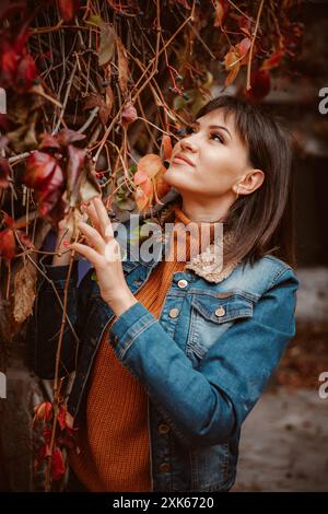 Eine Frau mit einer blauen Jeansjacke und einem orangefarbenen Pullover blickt nach oben und lächelt auf das bunte Laub um sie herum. Stockfoto