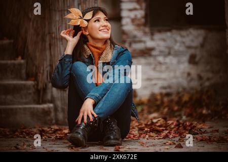 Eine Frau sitzt mit überkreuzten Beinen auf Steintreppen und trägt eine blaue Jeansjacke, Jeans und schwarze Stiefel. Herbstliche Blätter umgeben sie und sie smi Stockfoto