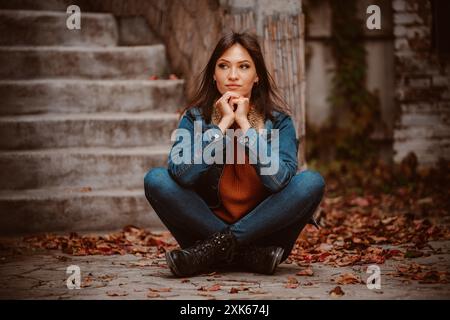 Eine junge Frau in Jeansjacke und Jeans sitzt mit überkreuzten Beinen auf Steintreppen, umgeben von gefallenen Blättern. Stockfoto