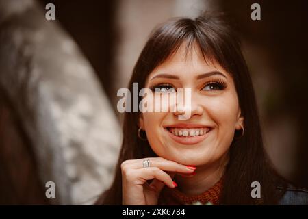 Eine junge Frau mit braunem Haar lächelt hell, lehnt ihre Hand auf ihr Kinn und blickt zur Seite. Stockfoto