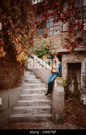 Eine junge Frau in einer Jeansjacke sitzt auf einer Steinmauer neben einer Betontreppe, die mit Herbstlaub bedeckt ist. Stockfoto