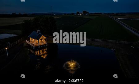 Ruhige Farm-Szene in der Abenddämmerung mit Einem gemütlichen Steinhaus, beleuchtet von sanfter Beleuchtung, reflektiert in Einem friedlichen Teich mit Einem Brunnen, umgeben von weitläufigen grünen Feldern Stockfoto