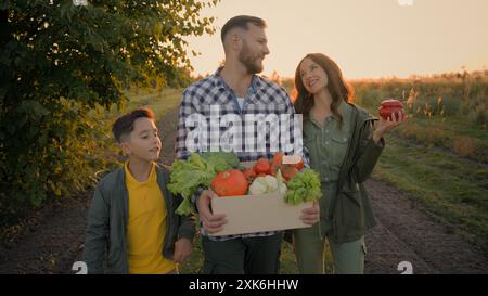 Glückliche kaukasische Familie Rückkehr nach der Landwirtschaft Arbeit ernten Mann männlicher Kerl Vater Tragen Kiste Gemüse kleines Kind Sohn Junge spazieren Frau Stockfoto