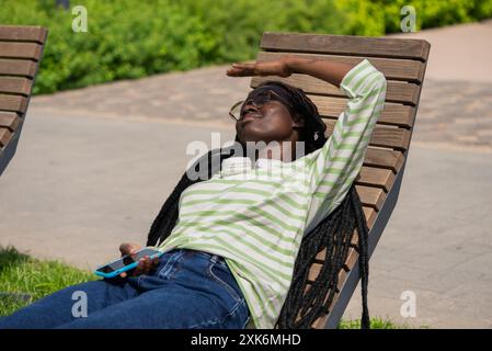 Afroamerikanerin mit Kopfhörern und Smartphone, die sich auf einer Holzbank entspannt und ihre Augen vor der Sonne im Park schützt. Das schwarze Mädchen versteckt sich vor den heißen sengenden Rochen. Stockfoto