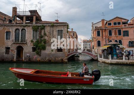 Venedig, Italien - 4. Juni 2024: Mann überquert einen Kanal mit rotem Boot. Campiello Mosca Straße. Die Migrantenkinder-Kunst von Banksy im Hintergrund. Stockfoto