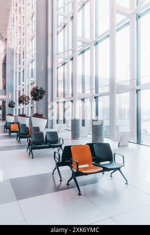 Vertikale Weitwinkelaufnahme eines modernen Flughafenwartebereichs mit großen Glasfenstern, Kunststoffstühlen mit schwarzen und orangefarbenen Sitzen und Innenpflanzen in W Stockfoto
