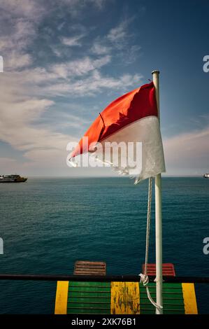 Indonesische Flagge über dem ruhigen Meer, Eine alte zerrissene indonesische Flagge weht im Wind vor einem ruhigen blauen Ozean Hintergrund. Boote schweben am Horizont. Stockfoto