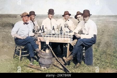 Sechs Männer, die draußen sitzen, Hüte tragen, spielen, trinken und Karten spielen, mit einem großen Gewehrgeschenk, um 1910. Stockfoto