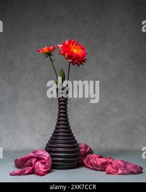 Stillleben mit zwei roten Blumen. Dahlien in einer dunklen Holzvase auf grauem Hintergrund. Wunderschöne, minimalistische Einrichtung mit Blumenmuster. Kopierbereich. Stockfoto