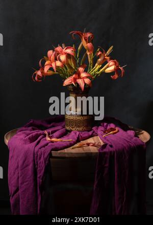 Ein Strauß orangener Lilien in einer grünen Vase, neben einem lila Schal. Grauer Hintergrund. Stillleben Stockfoto