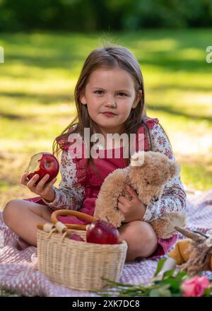 Porträt eines süßen kleinen Mädchens auf einem Picknick. Das Kind hält einen roten Apfel in der Hand und lächelt; in der Nähe befindet sich ein Obstkorb und ein Teddybär Stockfoto