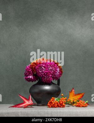 Stillleben mit Blumen und Beeren auf dunklem Hintergrund. Rosa, rote Dahlien in einer grauen Vase, Orangenbeeren, gelbe Blätter Stockfoto