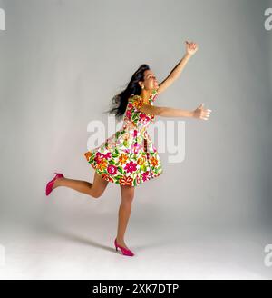 Fröhliche junge Frau in leuchtendem Blumenkleid und pinkfarbenen Absatzschuhen mitten im Sprung mit ausgestreckten Armen, lächelnd vor einem sauberen grauen Studiohintergrund. Stockfoto