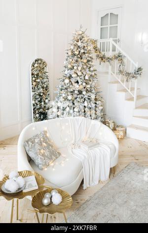 Modernes Wohnzimmer mit Treppe, einem in Silber und Gold dekorierten Weihnachtsbaum, weißem Sofa mit Kissen und Decke, Geschenke unter dem Baum. Die Stockfoto