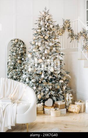 Elegantes Wohnzimmer mit einem weißen Sofa, einem wunderschön dekorierten Weihnachtsbaum mit goldenen Ornamenten und Geschenken. Ein Spiegel und eine Treppe runden das ab Stockfoto