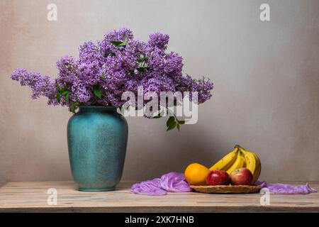 Ein großer Fliederstrauß in einer Vase auf hellgrauem Hintergrund, neben ihnen auf einem Teller sind Bananen, zwei rote Äpfel und eine Orange. Stillleben, viel Stockfoto