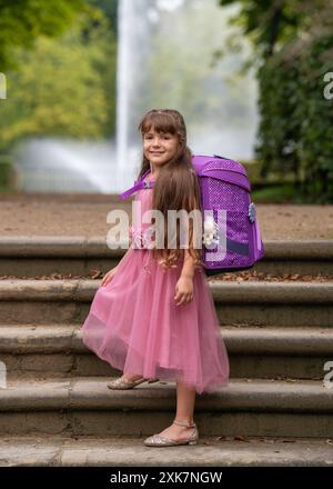 Ein kleines lächelndes Mädchen in einem rosa, schönen Kleid mit langen Haaren und Rucksack geht zur Schule, Foto auf den Stufen im Park. Vor dem Hintergrund Stockfoto