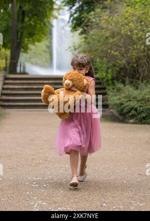 Ein kleines Mädchen in einem wunderschönen rosa Kleid mit langen Haaren läuft schnell und hält einen großen Teddybären in den Händen. Spiele im Freien. Freundschaft verschwommener Hintergrund Stockfoto