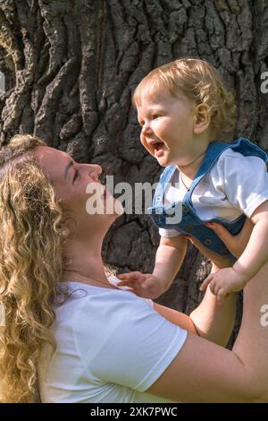 Porträt von Mutter und Sohn. Eine glückliche Mutter mit welligen Haaren hebt fröhlich ein kleines Baby mit welligen, blonden Haaren auf. Der Junge lacht fröhlich. Die Stockfoto