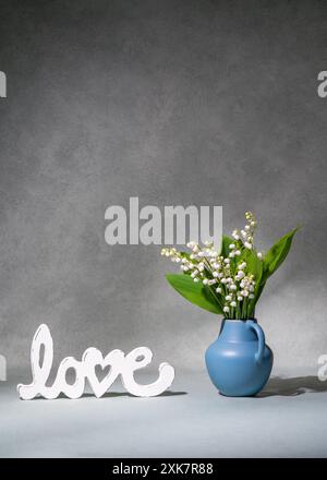 Ein kleiner Strauß weißer Blumen, Maiglöckchen aus dem Tal in einer blauen Vase neben der Aufschrift „Liebe“. Grauer Hintergrund. Platz für Text. Stillleben. Pos. Stockfoto